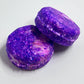 Two rounded shampoo bars with a purple and pink marbled pattern, placed on a light grey surface.