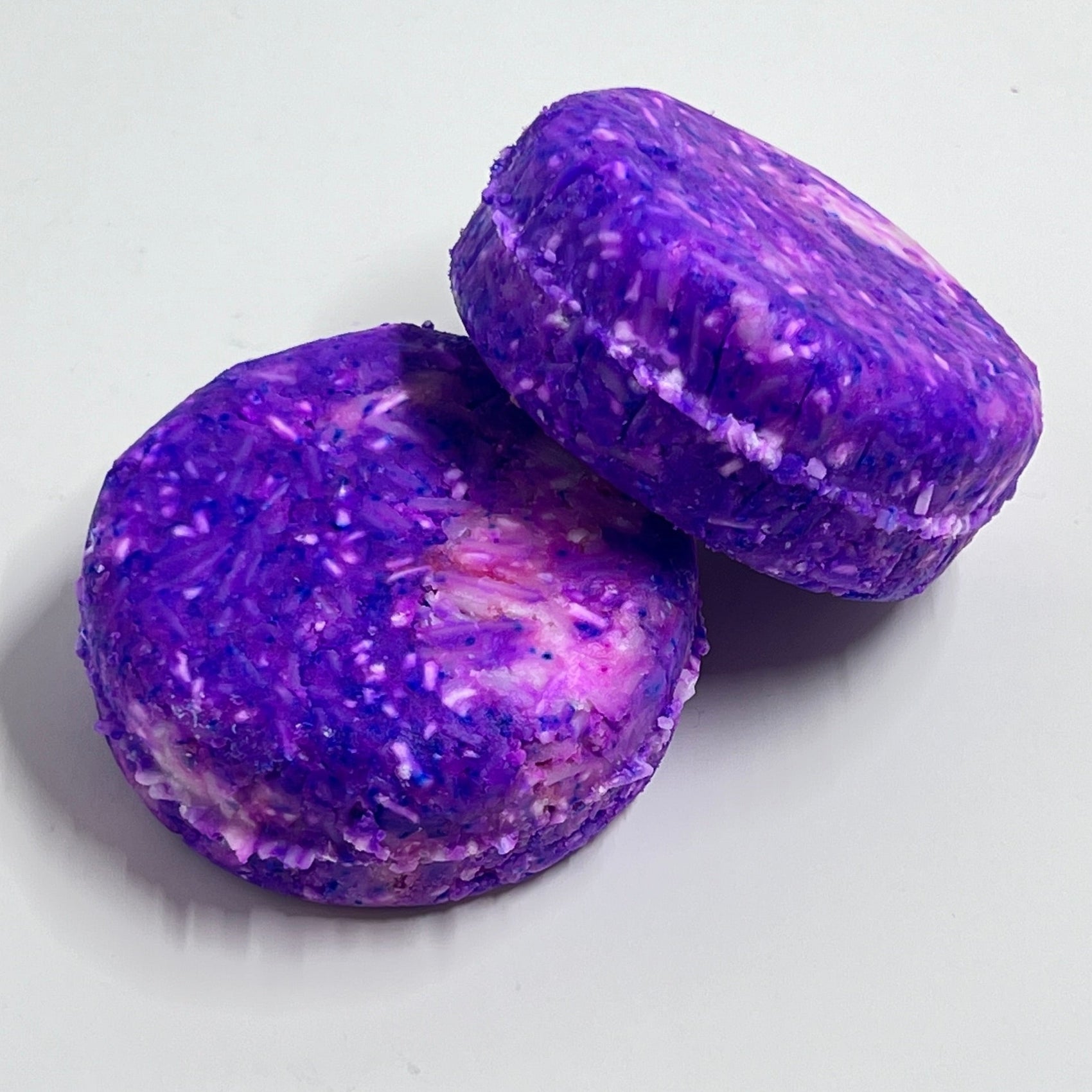 Two rounded shampoo bars with a purple and pink marbled pattern, placed on a light grey surface.