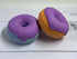 Two donut bath bombs with purple frosting one is blue and the other is orange