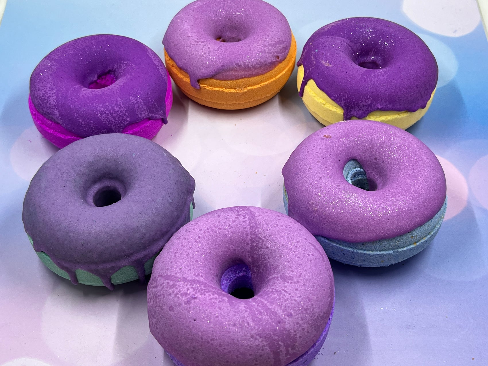 six donut bath bombs in a circle showing all colors of the rainbow. each have purple frosting and they are sitting on a multicolored background
