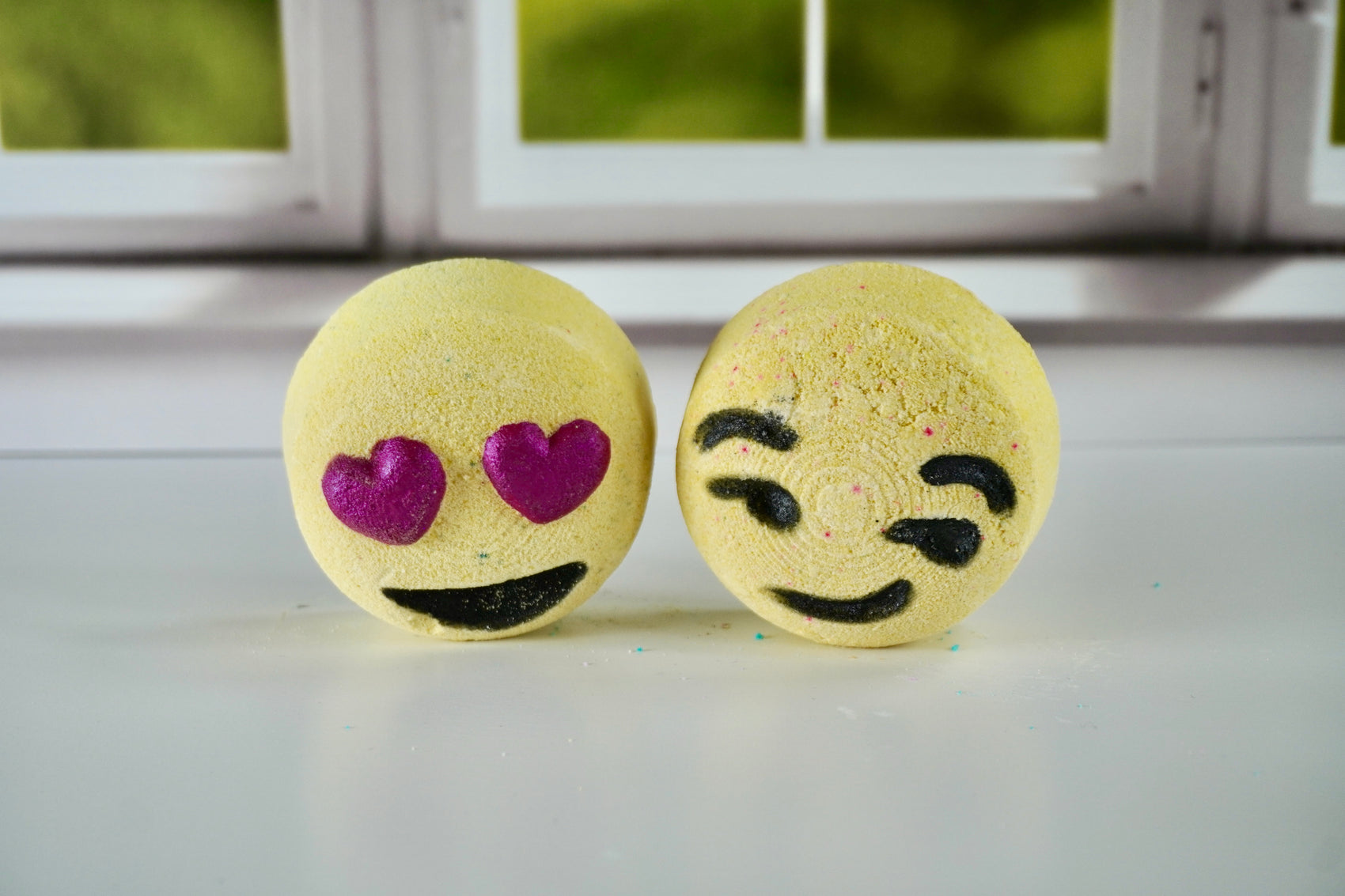 Two bath bombs with emoji faces on a white surface, one with heart eyes and the other with a sad face.