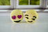 Two bath bombs with emoji faces on a white surface, one with heart eyes and the other with a sad face.
