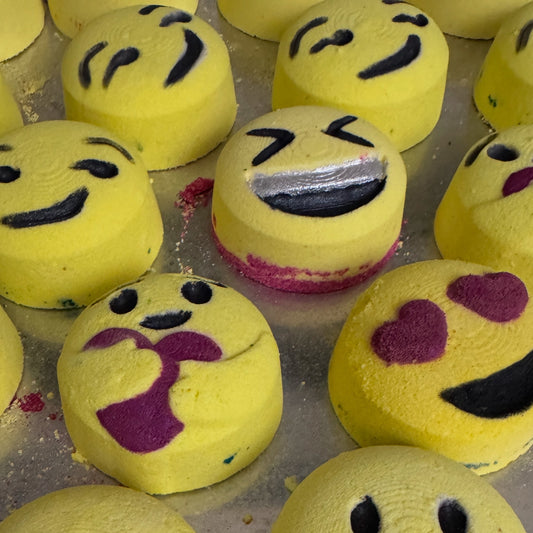 Yellow emoji-themed bath bombs with various facial expressions on a metal surface.