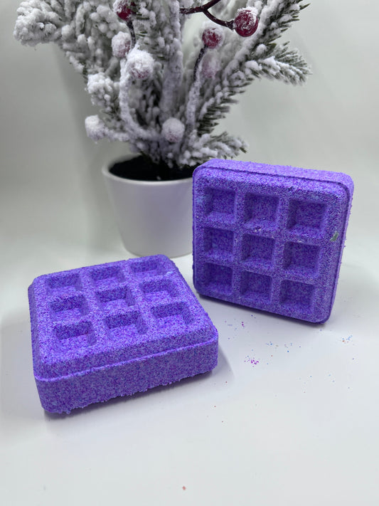 Two purple waffle bath bombs in front of a tree covered in snow and a white background