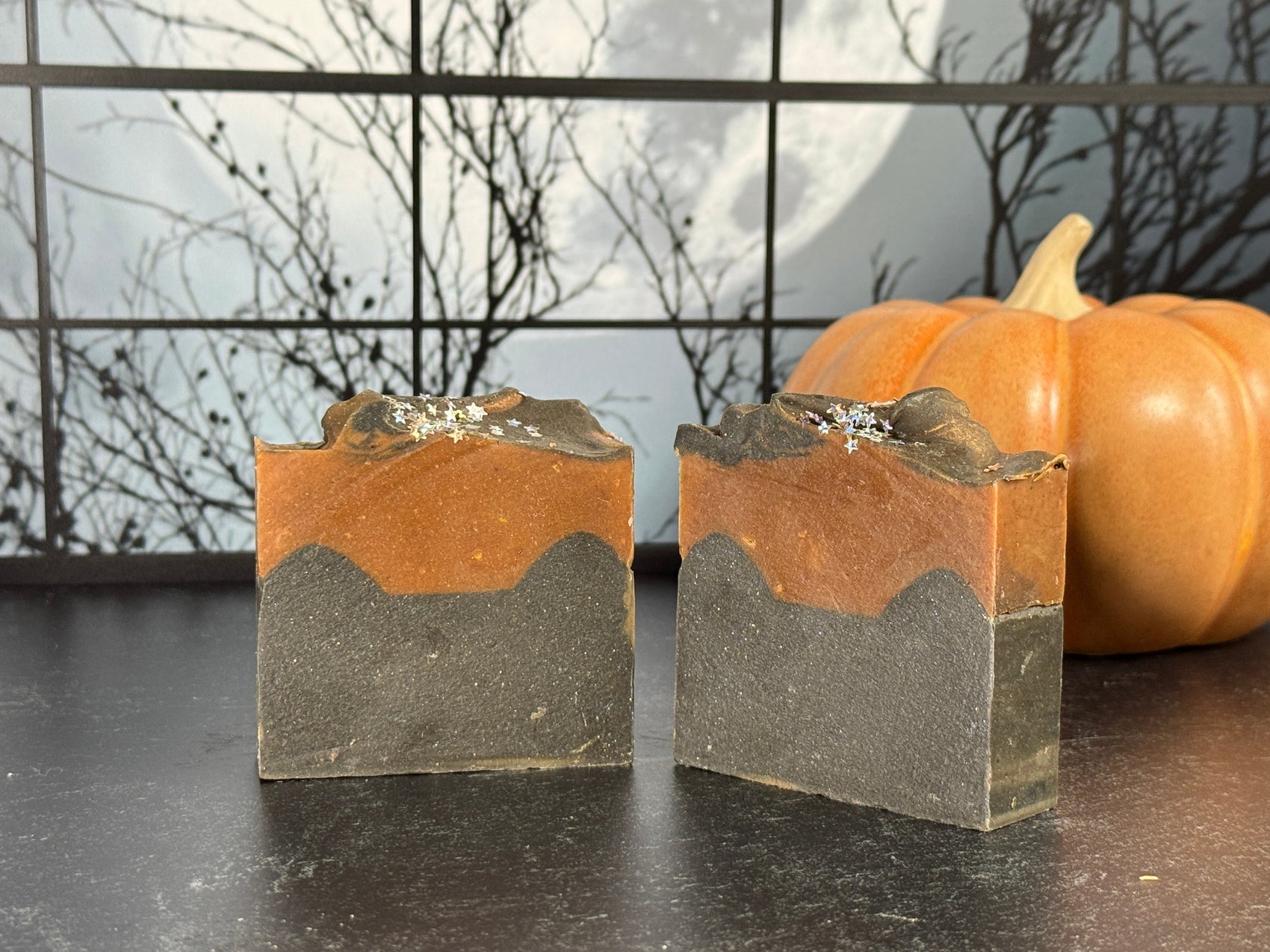 Two bars of orange and black layered artisan soap with glitter on top. A ceramic pumpkin is visible in the background, set against a spooky window view of bare tree branches and a full moon.