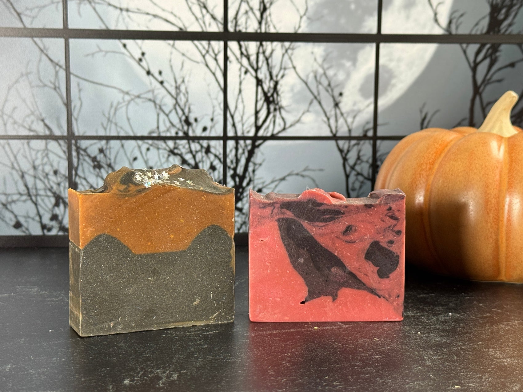 Two bars of Halloween-themed artisan soap: one orange and black layered, the other red and black marbled with a Crow shape. A ceramic pumpkin sits in the background against a spooky window scene with a full moon.