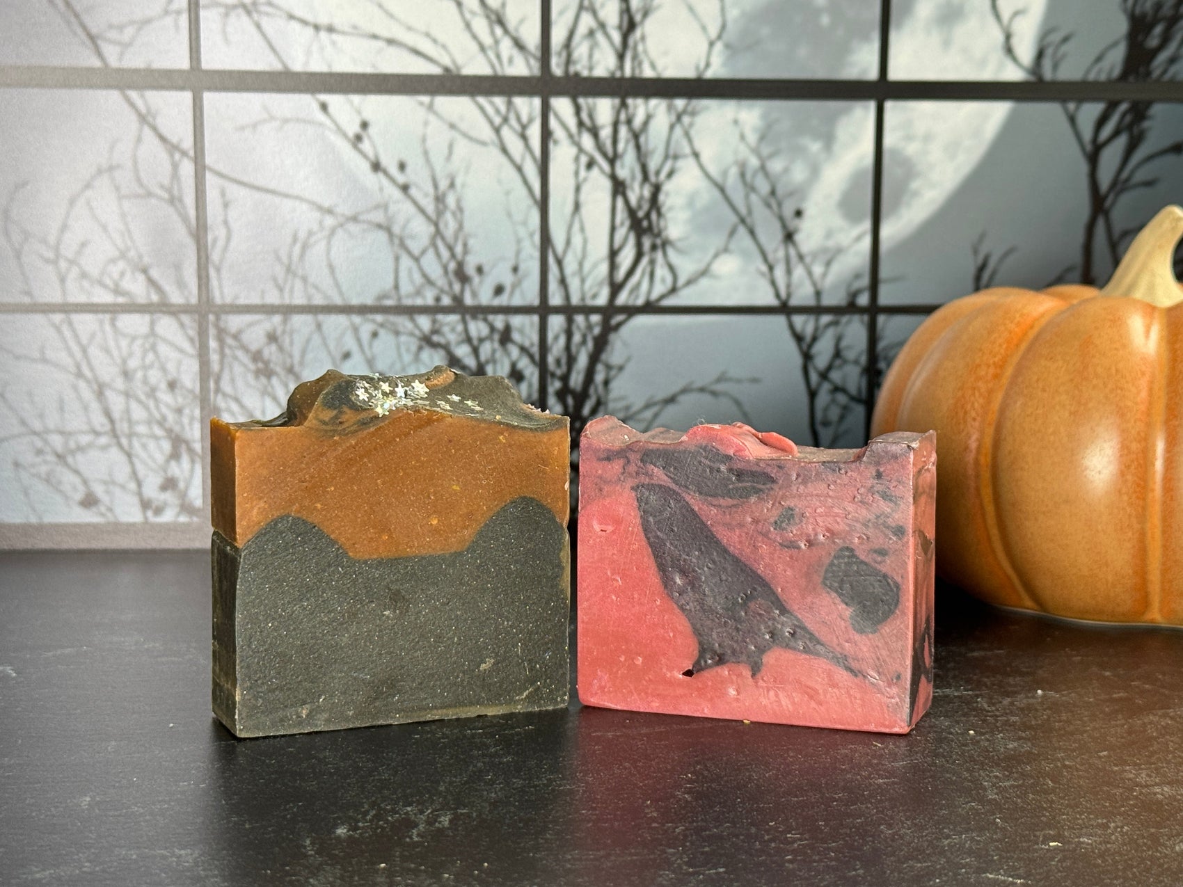 Two bars of Halloween-themed artisan soap: one orange and black layered, the other red and black marbled with a crow shape. A ceramic pumpkin sits in the background against a spooky window scene with a full moon.