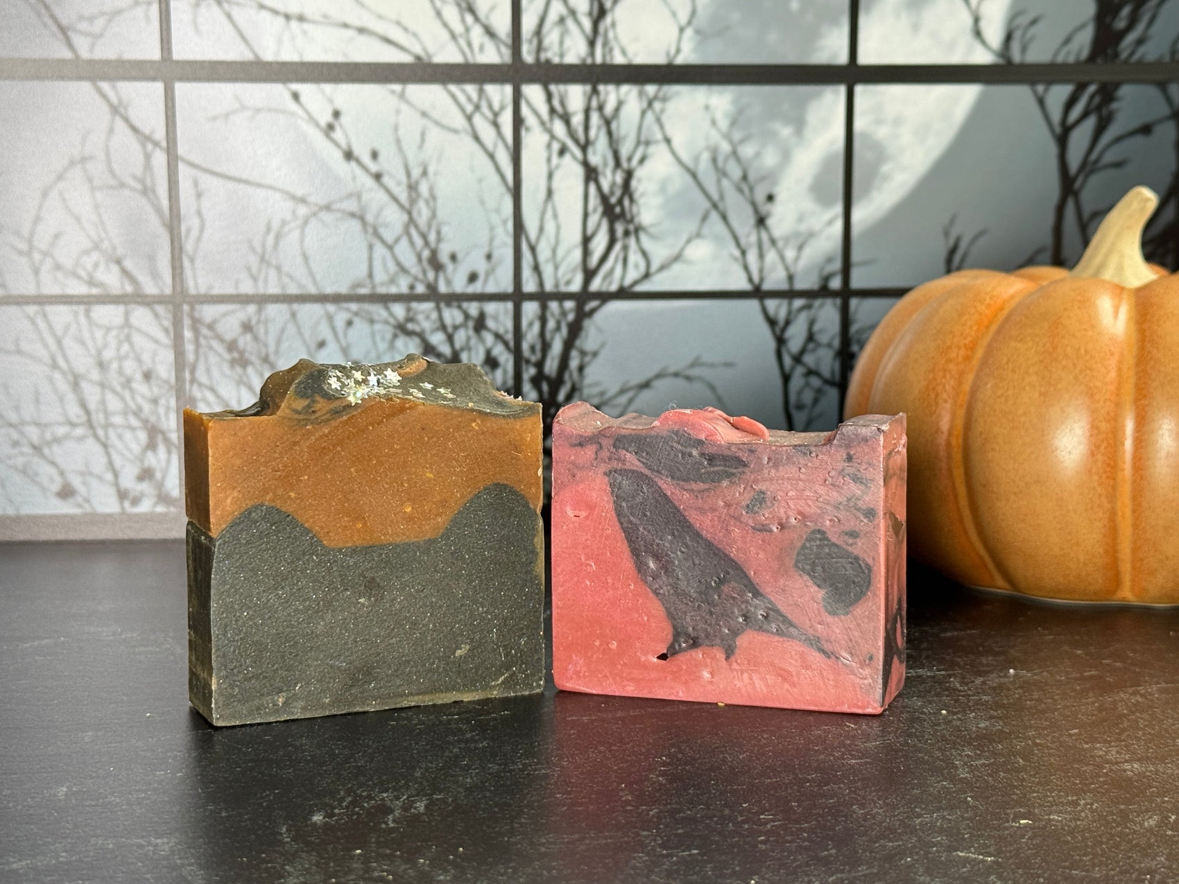 Two bars of Halloween-themed artisan soap: one orange and black layered, the other red and black marbled with a crow shape. A ceramic pumpkin sits in the background against a spooky window scene with a full moon.