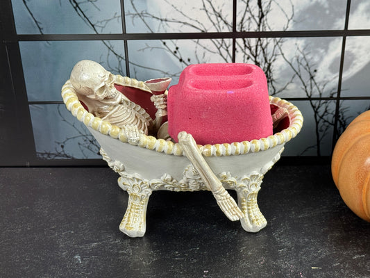 Pink Toaster bath bomb resting in a miniature white clawfoot bathtub with a relaxed skeleton, set against a spooky Halloween backdrop.