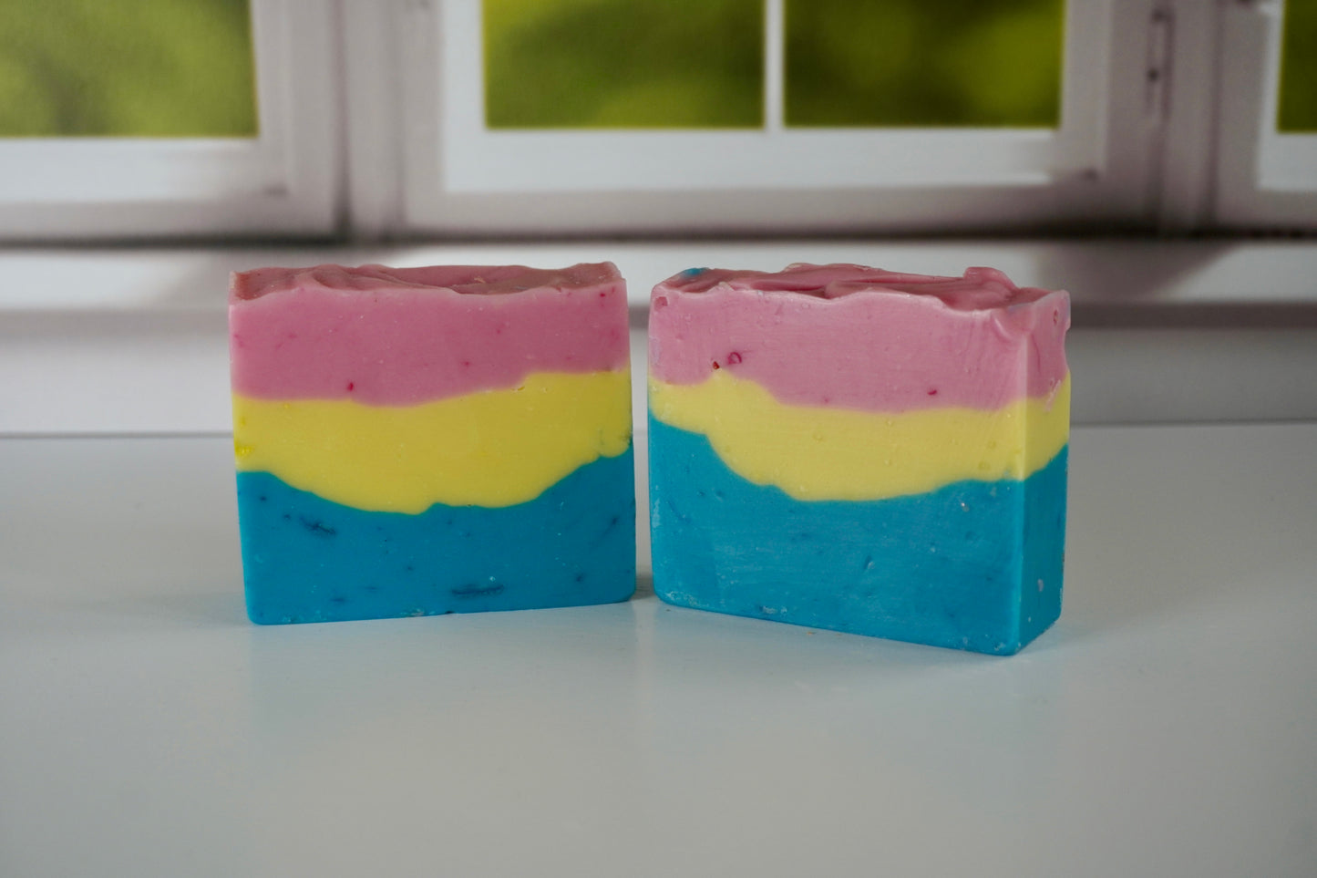 Two layered soap bars in pink, yellow, and blue on a white surface with a window in the background.