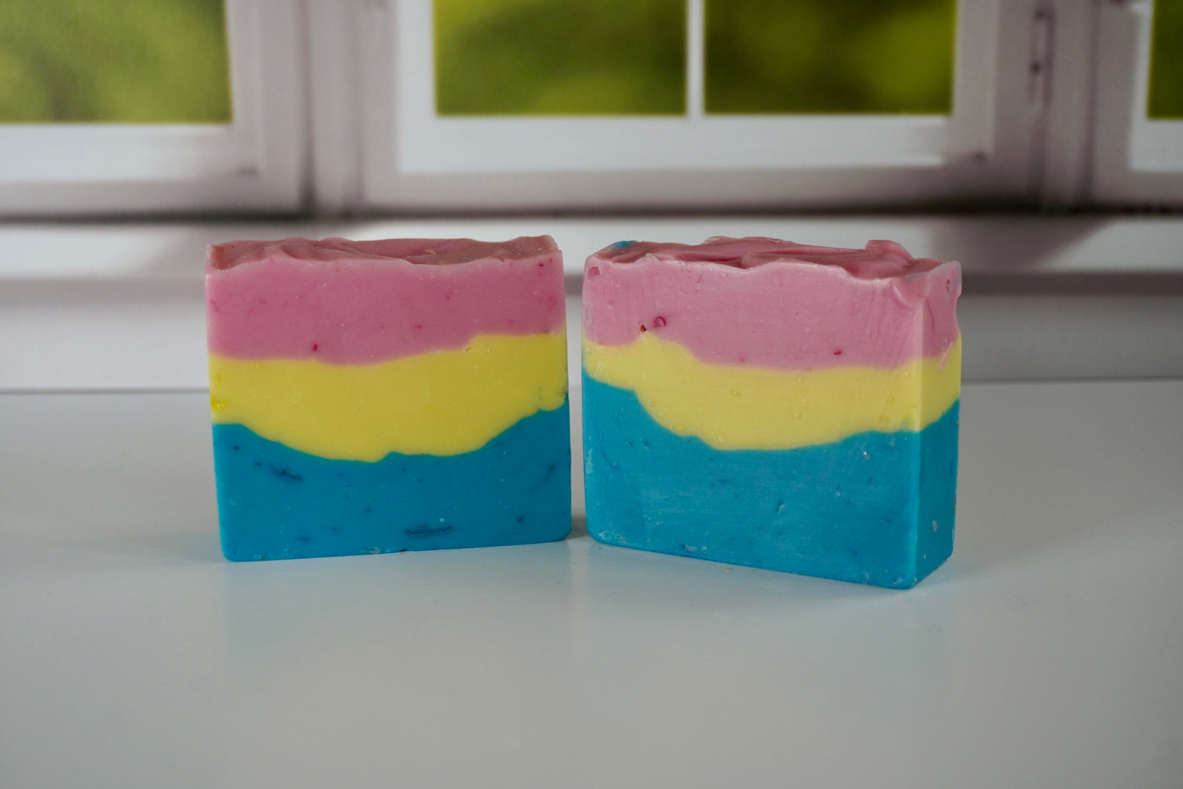 Two layered soap bars in pink, yellow, and blue on a white surface with a window in the background.