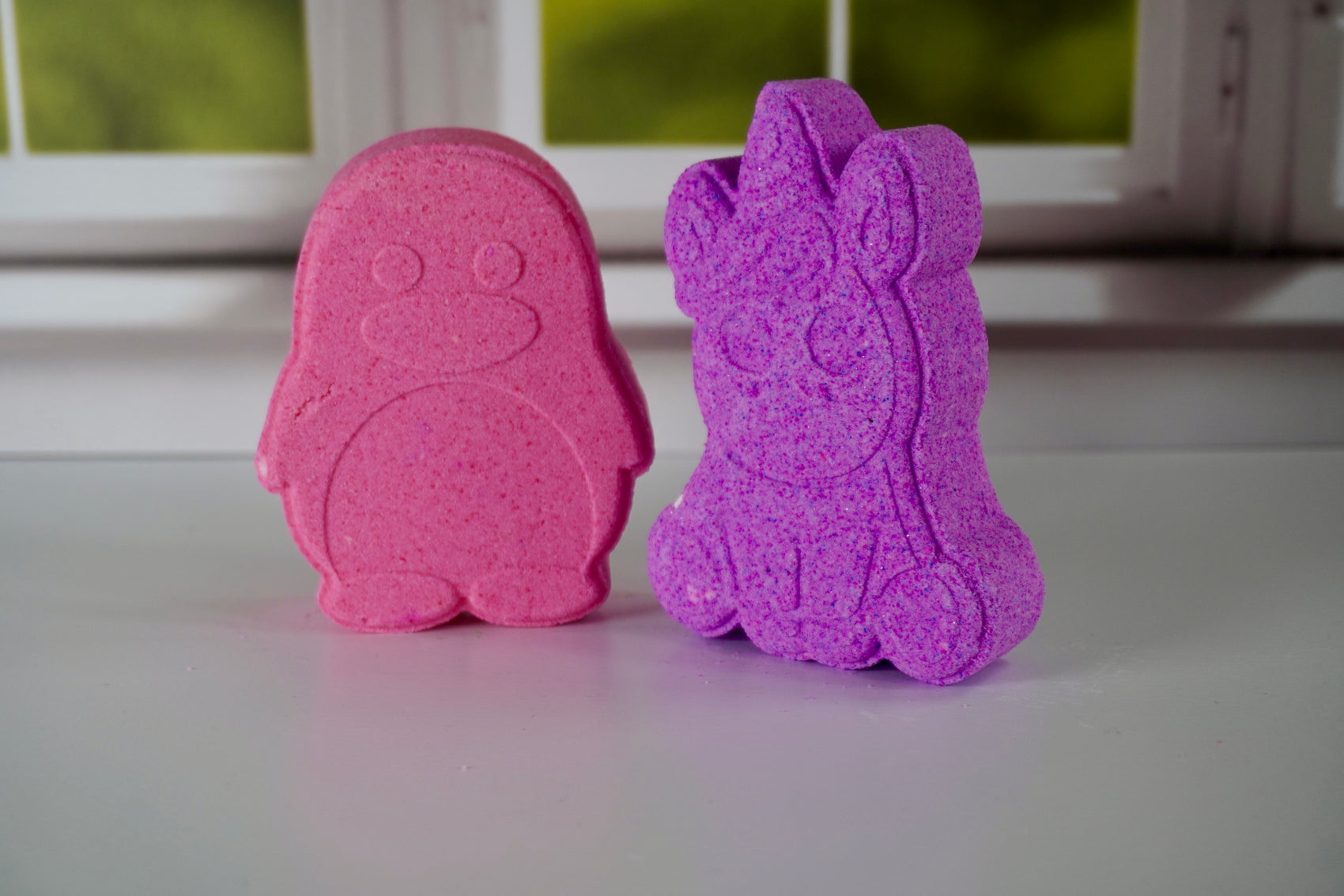 Pink and purple penguin and unicorn bath bombs on a white surface with a blurred background