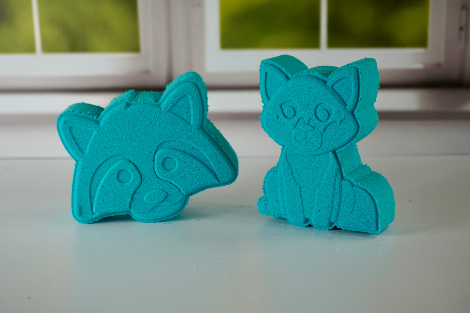 Two teal racoon and fox shaped bath bombs sitting side by side in front of a window