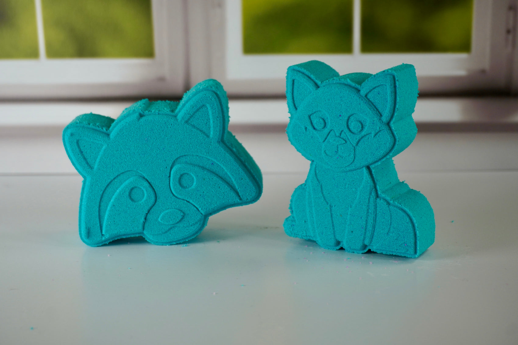 Two teal racoon and fox shaped bath bombs sitting side by side in front of a window
